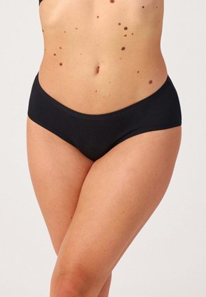 Żeński tors w czarnych majtkach o niskim stanie, widoczne znamiona na torsie, stojący na jednolitym jasnym tle.