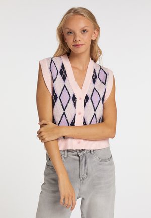 Jonge vrouw draagt een roze mouwloos vest met zwart en paars ruitpatroon en lichtgrijze spijkerbroek, staat met gekruiste armen.