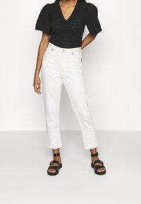 Černá texturovaná halenka s nařasenými rukávy, kombinovaná s bílými rovnými džíny a černými sand�ály na pásky. Pohled zepředu na jednoduchém pozadí.