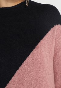 Svart och ljusrosa hoodie i grovstickad kvalitet. Texturerad tyg med ett diagonalt färgblockdesign. Har en liten metallaccent vid halslinningen.