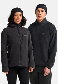 Due persone che indossano giacche in pile nere e berretti di lana neri abbinati, in piedi fianco a fianco su uno sfondo bianco.