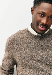 Brauner Strickpullover mit strukturiertem Muster und bunten Sprenkeln; getragen über einem weißen Hemd mit Kragen. Fokus auf den Hals- und Schulterbereich.