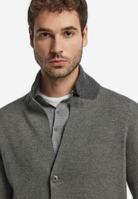 Grå cardigan med en texturerad krage, enkel knappstängning, bärs över en ljusgrå skjorta, vilket visar ett mjukt stickat material.