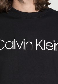 Černá mikina s bílým logem "Calvin Klein" na hrudi. Materiál je hladký s jemnou texturou a žebrovaným výstřihem.
