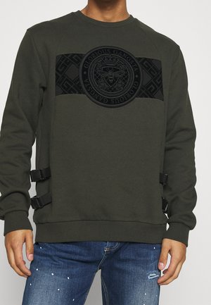 Oliwkowa zielona bluza z czarnym geometrycznym panelem i okrągłym emblemtem z pszczołą oraz napisem "Glorious Gangsta", noszona z podartymi dżinsami.