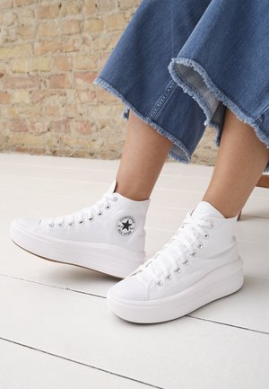 Personne portant des baskets montantes blanches Converse avec semelles compensées et un pantalon en jean bleu raccourci avec des ourlets effilochés.