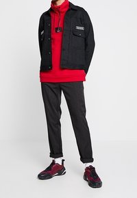 Czarna kurtka na guziki z logiem, czerwona bluza z kapturem, szare spodnie w paski, oraz czarno-czerwone sneakersy z białymi akcentami.