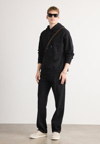Zwarte hoodie met een voorzak en trekkoorden, gecombineerd met zwarte broeken en crèmekleurige sneakers. Model draagt zwarte zonnebril en een bruine schoudertas.