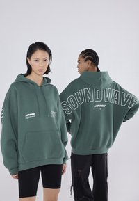 Πράσινη oversized κουκούλα με μπροστινή τσέπη, featuring λευκό κείμενο "SOUNDWAVE" και λογότυπο της μάρκας. Το μοντέλο φοράει μαύρα σορτς και σκισμένα τζιν.