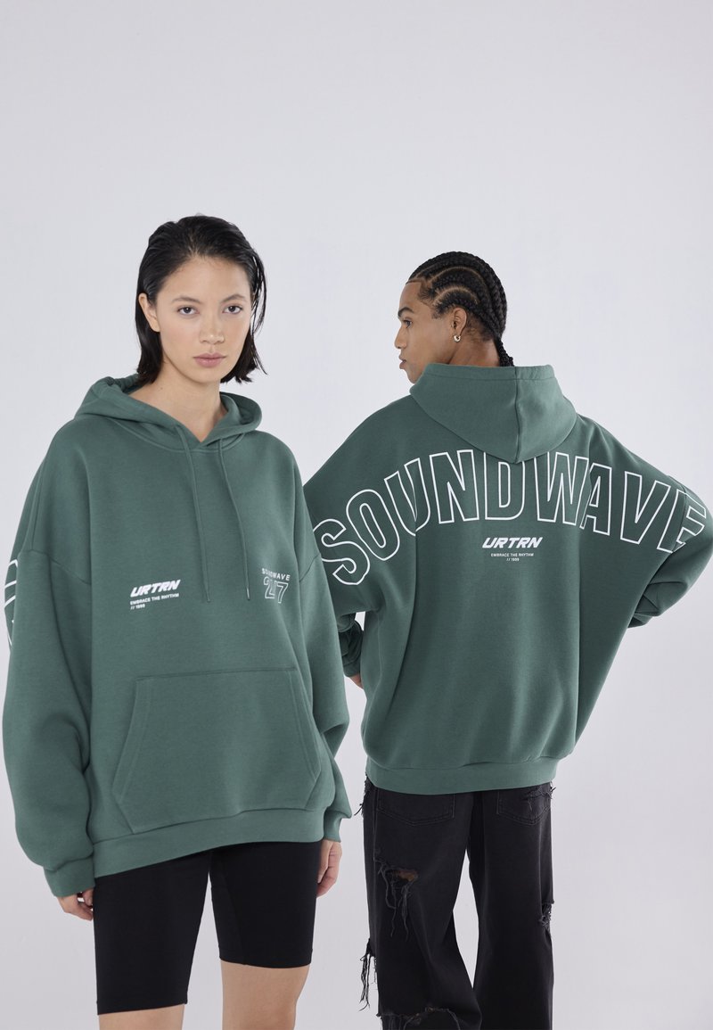 Πράσινη oversized κουκούλα με μπροστινή τσέπη, featuring λευκό κείμενο "SOUNDWAVE" και λογότυπο της μάρκας. Το μοντέλο φοράει μαύρα σορτς και σκισμένα τζιν.