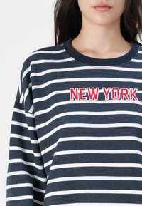 Marinefarget hettegenser med hvite horisontale striper, rund halslinning og rød brodert "NEW YORK" tekst på brystet.