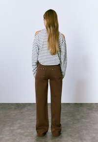 Top de manga larga a rayas gris y blanco con hombros descubiertos, combinado con pantalones de pierna ancha marrones que tienen bolsillos traseros y tachuelas decorativas.