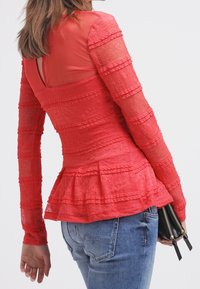 Top peplum rojo con tela texturizada, mangas largas y volante en capas en la cintura, combinado con jeans de mezclilla azules y un bolso negro pequeño.