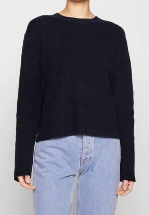 Osoba w czarnym swetrze z długim rękawem i jasnoniebieskich jeansach z wysokim stanem, stojąca na jednolitym tle.