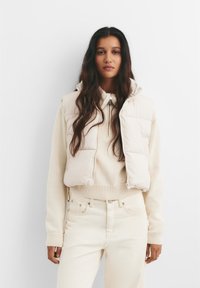 Bež prsluk s kapuljačom, na zatvaranje sa zipom i kvadratnim šavovima, nosi se preko krem pletenog džempera i odgovarajućih svijetlih hlača.