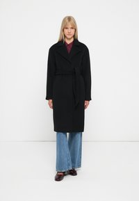 Cappotto di lana nera con cintura, dotato di collo a rever e maniche lunghe, abbinato a jeans azzurri a gamba larga e scarpe scure.