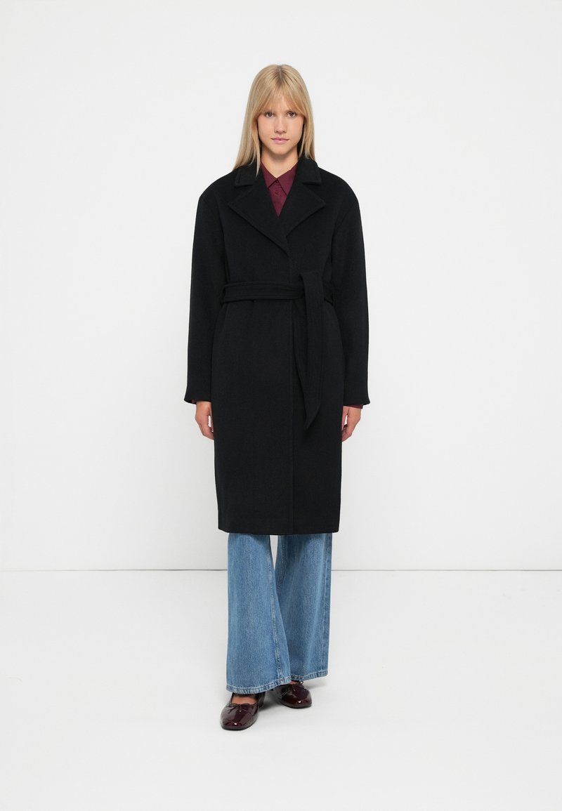 Abrigo de lana negro con cinturón, con cuello de muesca y mangas largas, combinado con vaqueros de pierna ancha de color azul claro y zapatos oscuros.