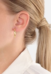 Närbild av ett öra med en vriden guldring som örhänge, blont hår i hästsvans och iförd en vit skjorta med krage.