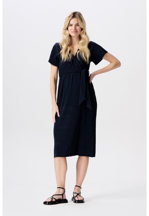 Robe portefeuille bleu marine, longueur genou, avec des manches courtes et un lien à la taille, présentant une texture côtelée. Associée à des sandales plates noires.