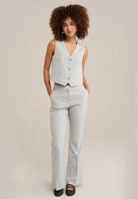 Completo grigio chiaro a tre pezzi con gilet senza maniche e bottoni, pantaloni su misura e scarpe basse. Tessuto liscio e design aderente.