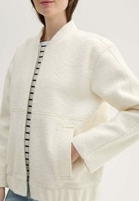 Weiße Fleecejacke mit Reißverschluss, geripptem Saum und seitlichen Taschen. Trägt man über einem schwarz-weiß gestreiften Shirt. Weicher Stoff, lockere Passform.