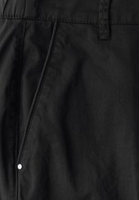 Svarta byxor i tyg med en slät yta, med sidofickor och synlig vit tryckknapp. Säkrade med en sydd midjeband.