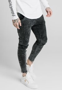Svarta, urblekta skinny jeans med sliten textur och subtila whiskeringdetaljer, kombinerade med vita höga sneakers med gulddetaljer.