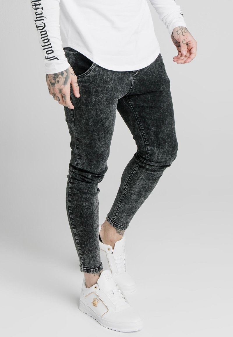 Svarta, urblekta skinny jeans med sliten textur och subtila whiskeringdetaljer, kombinerade med vita höga sneakers med gulddetaljer.