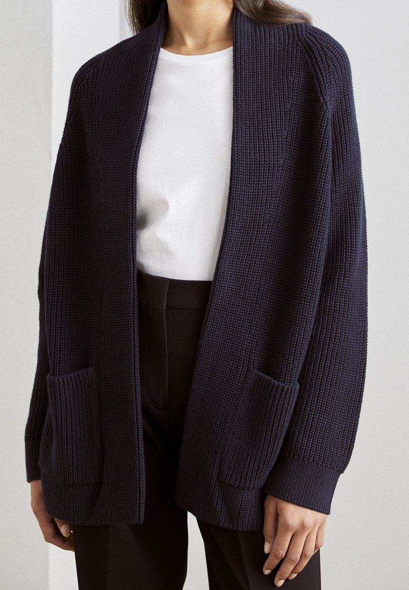 Cardigan en maille marine avec manches longues, devant ouvert, texture côtelée et deux poches avant. Porté par-dessus une chemise blanche et un pantalon noir.