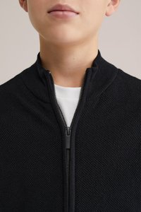 Svart zip-up stickad jacka med hög krage. Har ett strukturerat tyg och en silverfärgad dragkedja framtill, lager-på-lager över en vit skjorta.