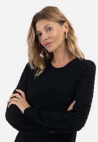 Fekete, textúrált pulóver, lekerekített nyakkivágással és hosszú ujjakkal, kiemelkedő mintázattal. Gyöngy fülbevalóval viselve.