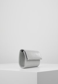 Petite pochette pailletée argentée avec rabat, posée à la verticale sur un piédestal blanc devant un fond gris uni.