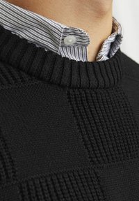 Melnā adīta džemperis ar ribotu kakla izgriezumu un teksturētām rakstainām detaļām. Zem džempera ir redzama svītraina krekla apkakle.