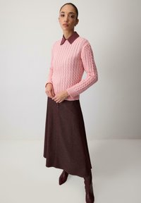 Žena v ružovom svetri s vrkočovým vzorom, bordovej košeli s golierom, tmavohnedej sukni a bordových čižmách stojaca na jednofarebnom pozadí.