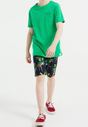 Niño caminando con una camiseta verde brillante, pantalones cortos azul marino con estampado de aves tropicales y zapatillas rojas con cordones blancos.