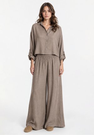 Donna che indossa una camicia taupe larga con bottoni e maniche arrotolate e pantaloni larghi abbinati, in piedi davanti a uno sfondo semplice.