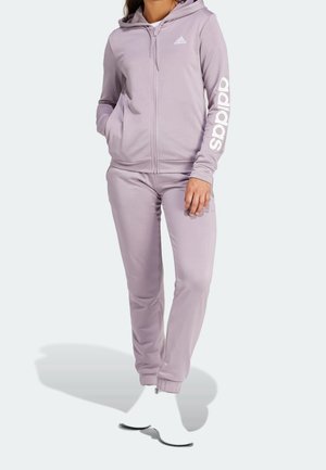 Kobieta w jasnofioletowym dresie Adidas z bluzą na suwak z kapturem i dopasowanymi spodniami, z rękami w kieszeniach, stojąca na białym tle.