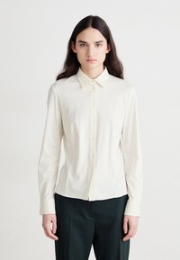 Langärmliges crèmefarbenes Hemd mit Kragen, Vorderknöpfen und tailliertem Design. Kombiniert mit dunkelgrünen Hosen. Sanfte Textur.