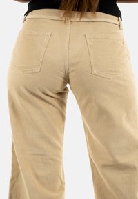 Beige corduroy broek met verticale ribbelstructuur, achterzakken en een taillebanddetail. De stof lijkt zacht en duurzaam.