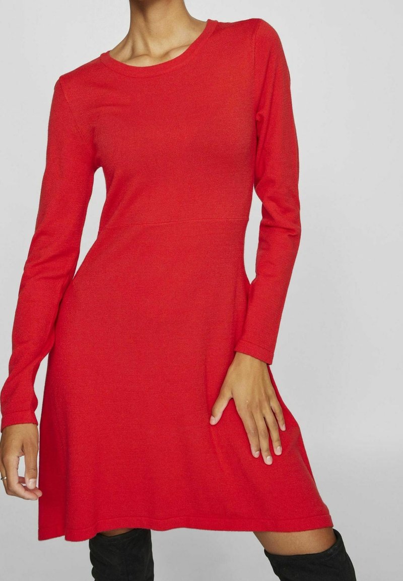 Femme portant une robe rouge à manches longues et longueur genoux associée à des bottes noires montantes sur un fond uni.