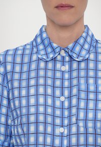 Personne portant une chemise à manches longues à carreaux bleus et blancs avec un col arrondi et une poche poitrine, sur un fond clair uni.