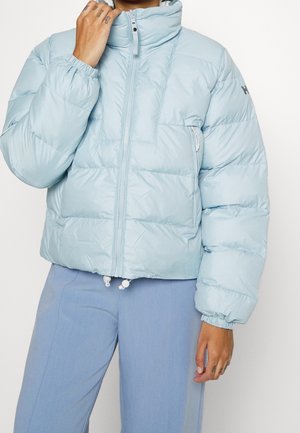 Personne portant une doudoune bleu clair avec fermeture éclair et poignets élastiques, assortie à un pantalon bleu clair, main touchant le col.