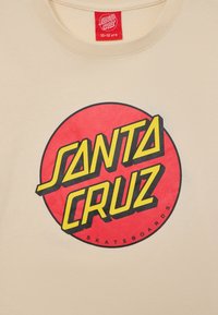 Šviesiai ruda medvilninė marškinėliai su ryškiu raudonu apskritimu, kuriame užrašyta "SANTA CRUZ" geltona ir juoda spalvomis, o žemiau - "SKATEBOARDS" juoda spalva.