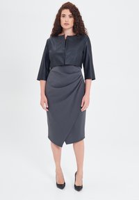 Giacca cropped in ecopelle nera con maniche tre quarti abbinata a un elegante abito grigio lungo fino al ginocchio con un design a drappeggio.