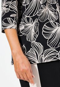 Schwarzes Oberteil mit weißen Blumenmustern und Dreiviertelärmeln, mit glatter Textur und einem Saum, der auf der Hüfte endet.
