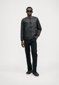 Giacca nera trapuntata con colletto rotondo e chiusure a nodo, indossata sopra una maglia verde e pantaloni neri. Modella con occhiali da sole neri e scarpe.