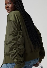 Olive green bomber jacket in shiny nylon, featuring gathered sleeves, ribbed cuffs, and hem. Paired with light blue jeans.
