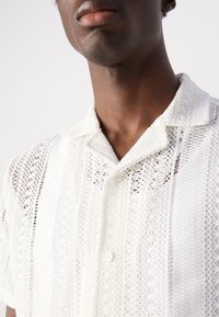 Cămașă albă cu mânecă scurtă, cu un design croșetat, care prezintă un guler cu nasturi și o textură modelată cu detalii verticale din tricot.
