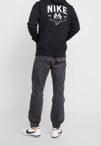 Svart sweatshirt med vit "NIKE"-logotyp och grafik på baksidan. Grå jeans med elastiska muddar, kombinerat med vita och svarta sneakers.