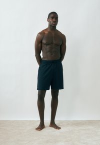 Μυώδης άνδρας στέκεται ξυπόλητος μπροστά σε έναν απλό ανοιχτόχρωμο τοίχο, φοράει σκούρα ριγέ σορτς μέχρι το γόνατο με τα χέρια πίσω από την πλάτη.